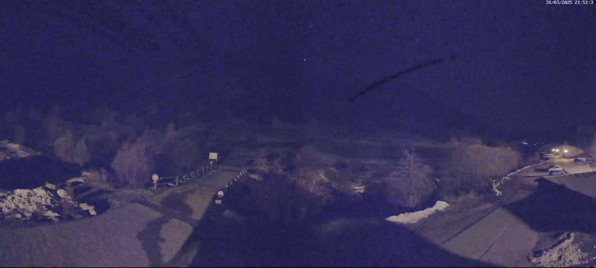 A nighttime, high-angle view shows a road with a few cars, some snow on the ground and bushes, and a dark sky with a plane's contrail.