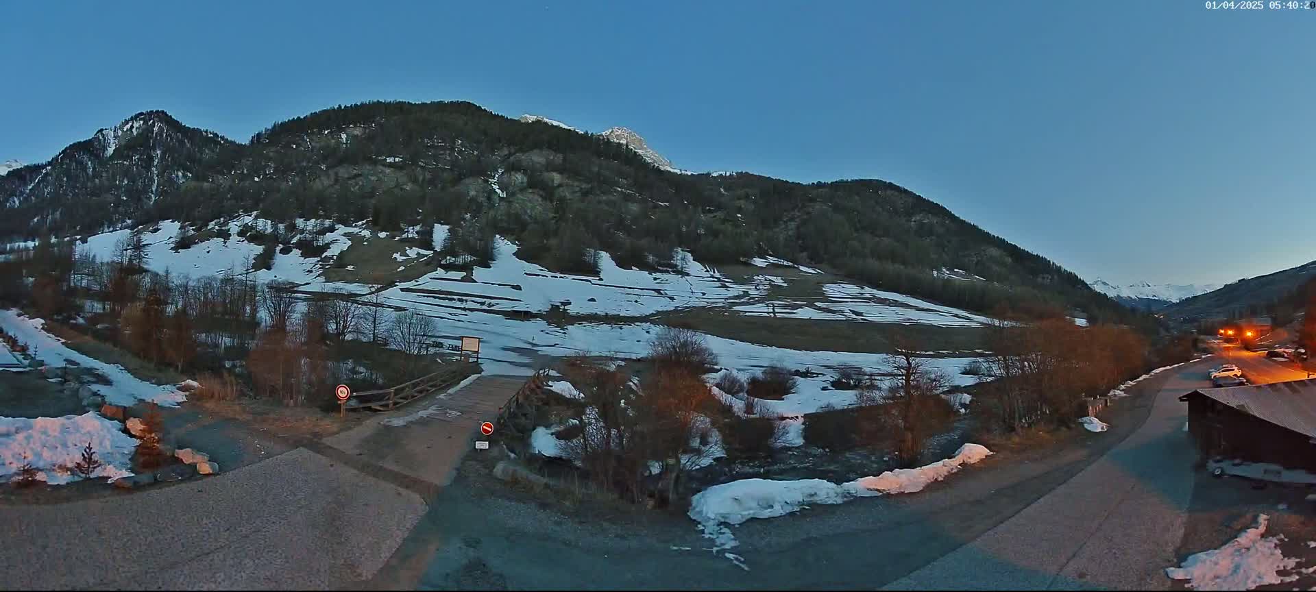 A snow-dusted mountain valley at twilight, with a small bridge over a stream and a road leading to a small building.