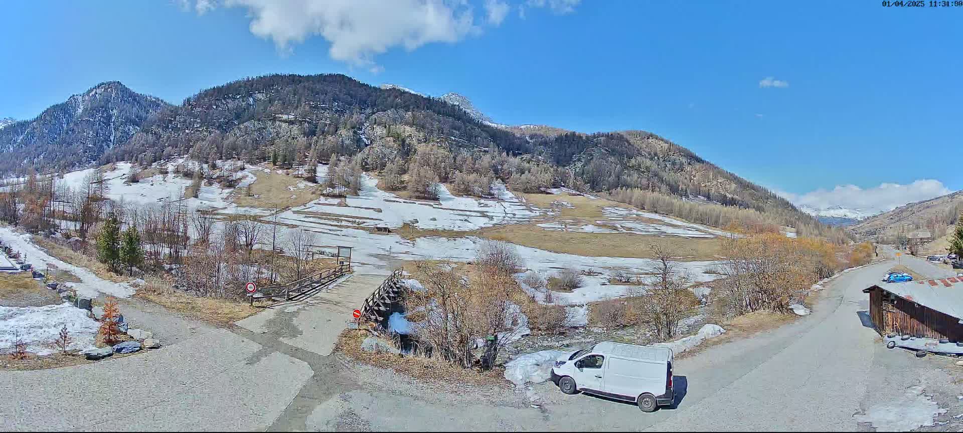 A mostly sunny day reveals a mountainous landscape with patches of snow and ice near a small bridge and road; a white van is parked nearby.