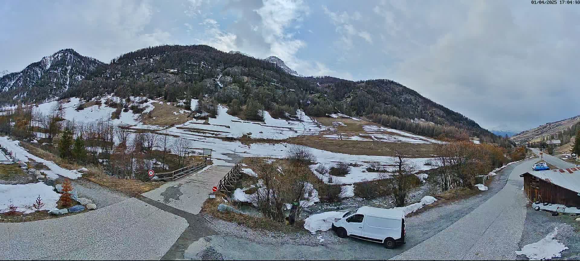 A mountain valley scene under a cloudy sky shows patchy snow on the ground and hillsides, a small wooden bridge over a stream, and a white van parked near a road.