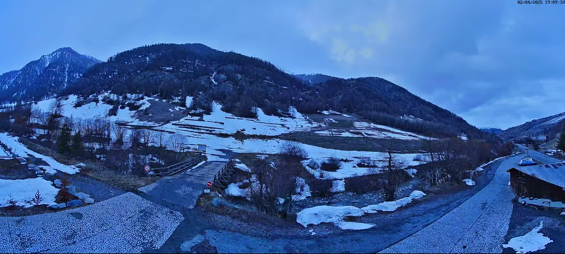 A snow-dusted mountain valley road leads to a small wooden bridge, with patchy snow covering the ground and hillsides under a twilight sky.