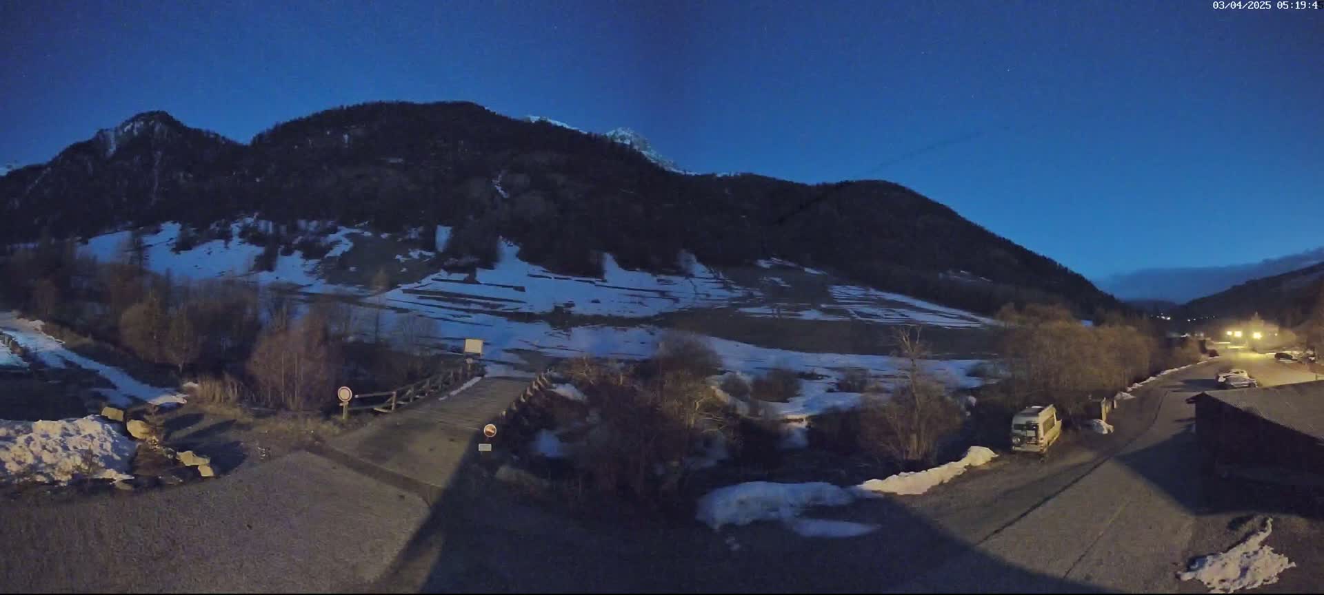 A nighttime view of a snow-dusted mountain valley with a small bridge and a few vehicles parked near a building under a clear, dark blue sky.