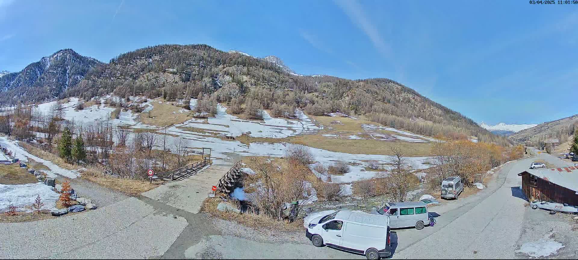 A sunny mountain valley scene with patches of snow on the ground, a few parked vans, and a small wooden building.
