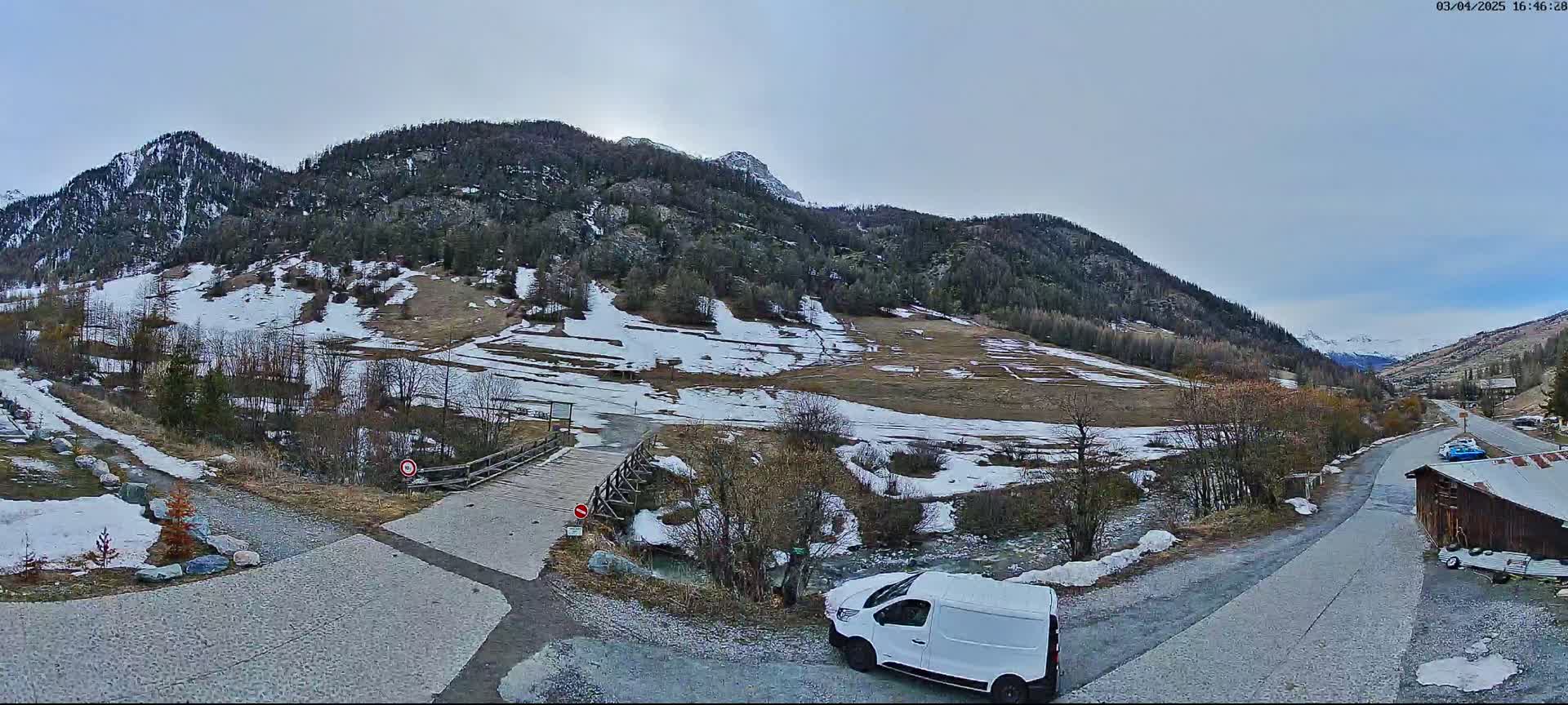 A white van is parked near a small wooden bridge crossing a stream in a valley, with snow patches on the ground and mountains in the background under an overcast sky.
