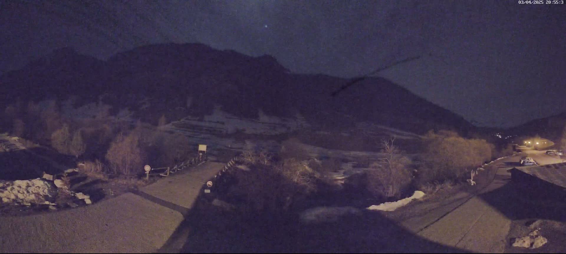 A nighttime view of a snow-dusted mountainside, a road, and some sparse vegetation under a dark sky.