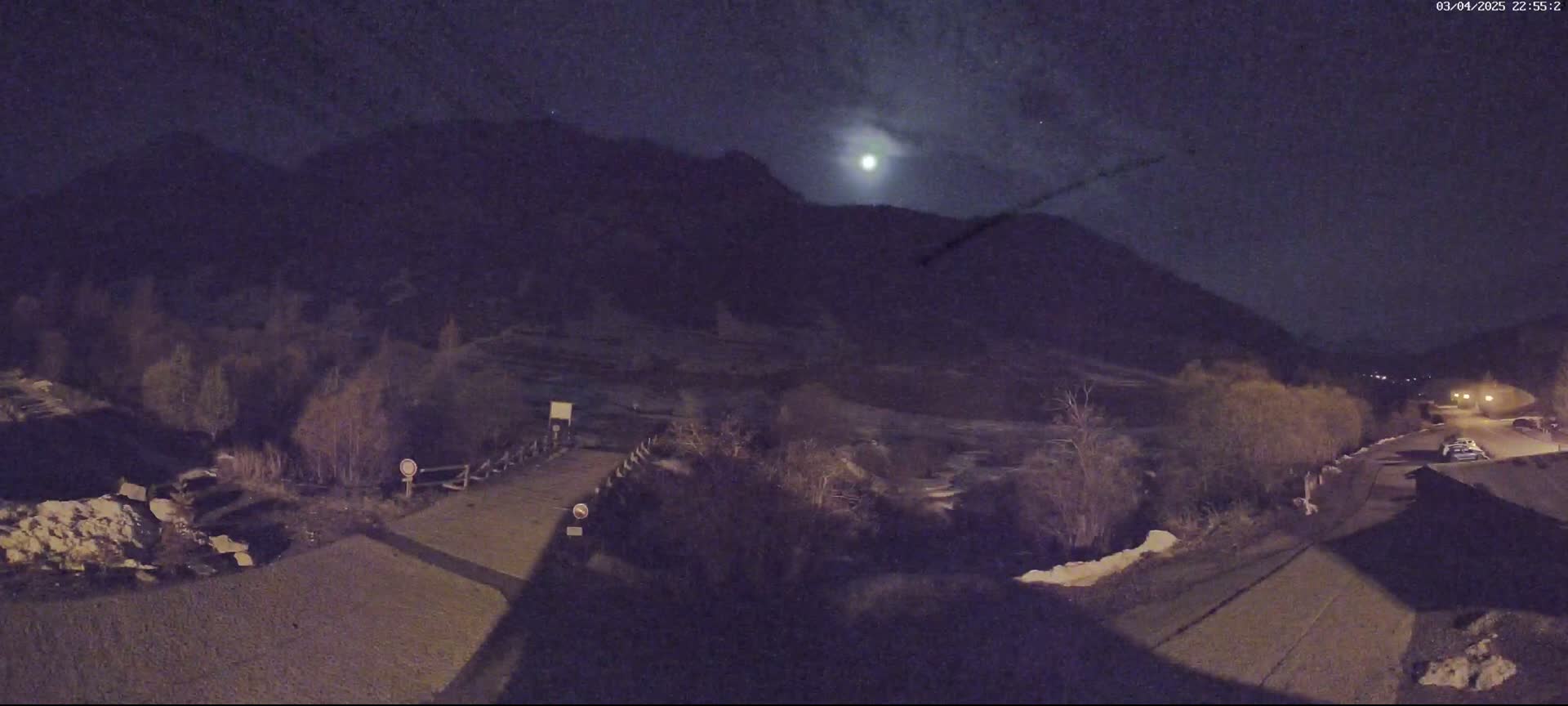 A nighttime view of a moonlit mountain landscape with a road leading to a small bridge and some buildings, featuring bare trees and patches of snow.