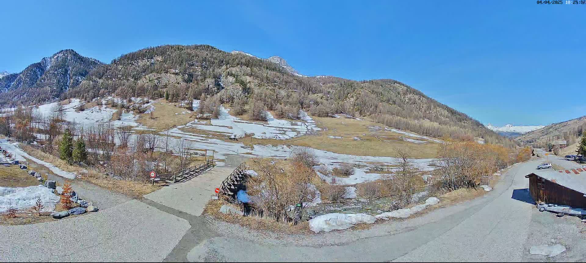 A sunny mountain valley scene with patchy snow on the ground and trees budding in early spring.