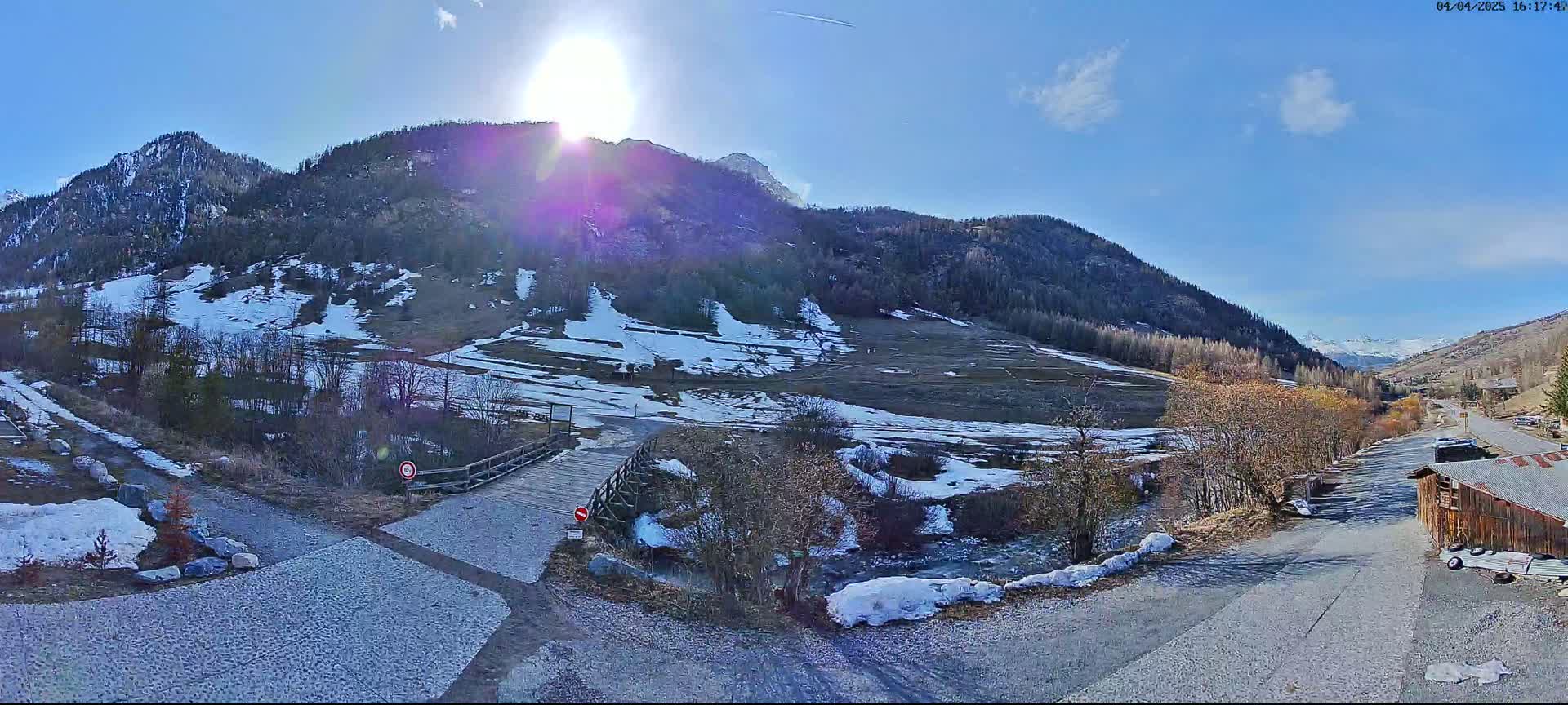 A sunny day in a mountainous region shows a small wooden bridge over a partially frozen stream, with patches of snow on the ground and hillsides.