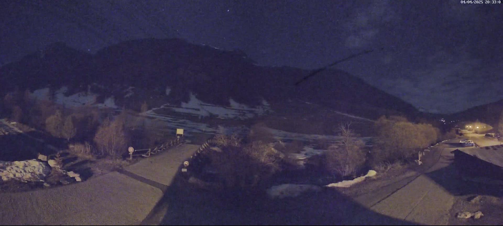 A nighttime view of a snow-dusted mountain valley, with a road leading to a small settlement under a dark, clear sky.