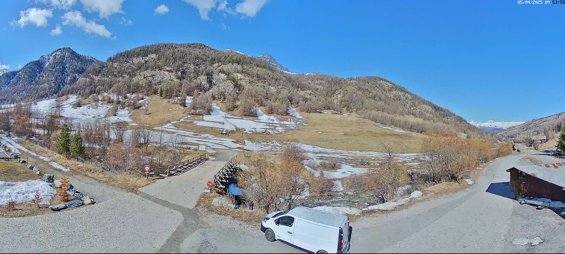 A white van is parked near a small bridge over a stream in a mountainous valley under a mostly sunny sky with patches of snow on the ground and hillsides.