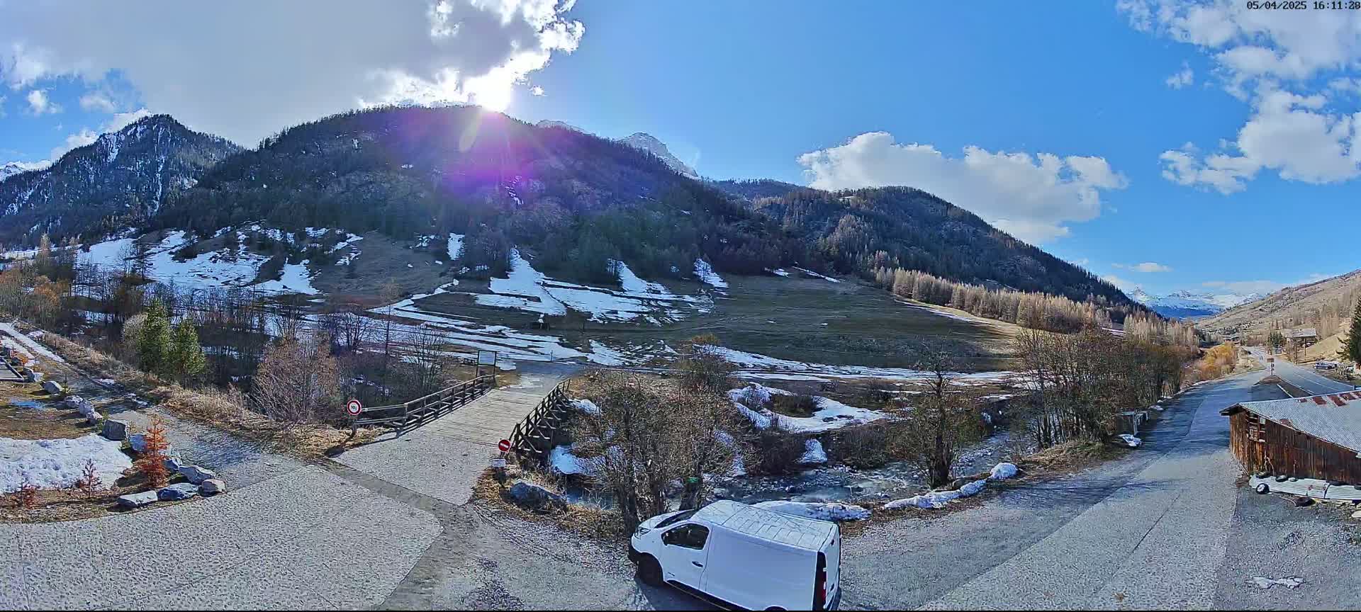 A sunny day in a mountainous area shows a valley with patchy snow, a small stream, a bridge, a white van, and a road leading to a building.