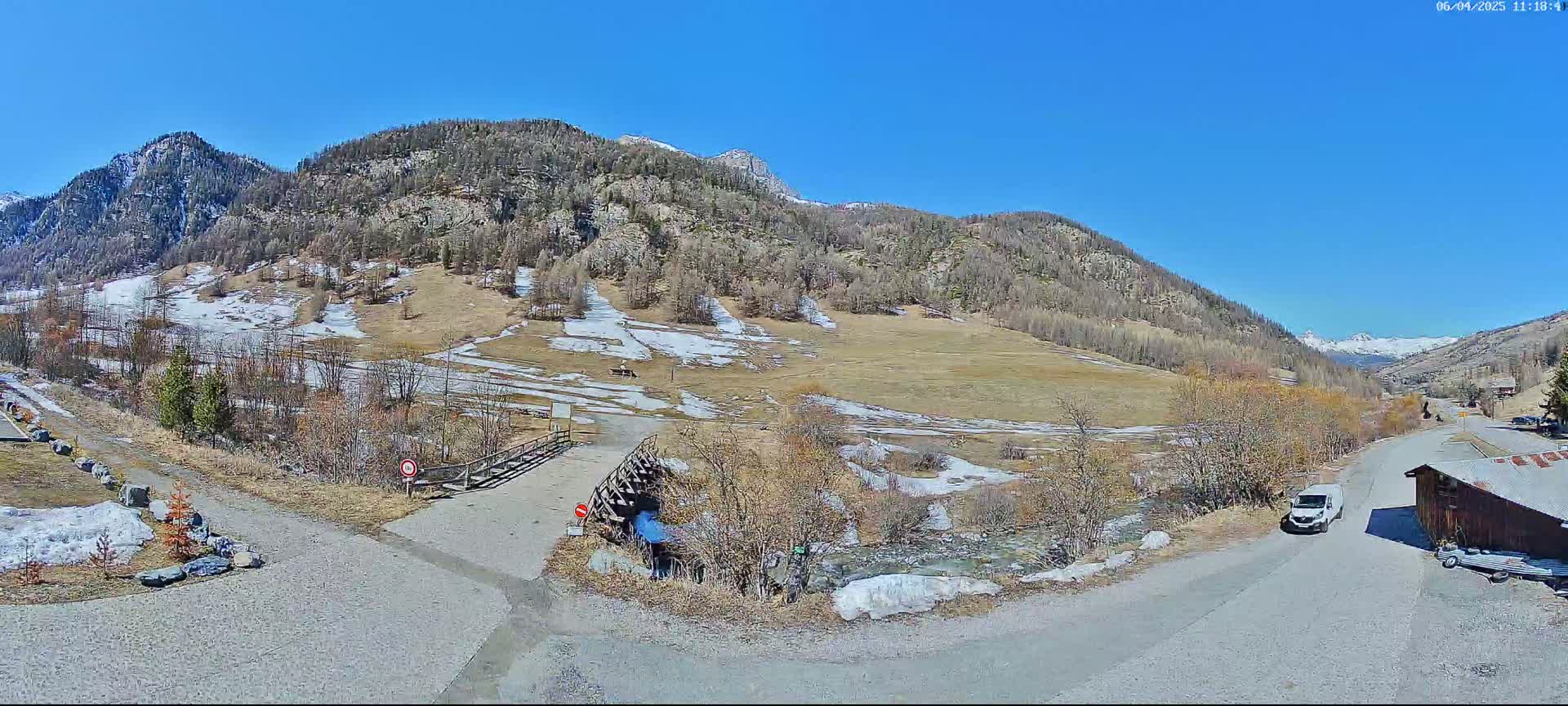 A mountain valley scene under a clear blue sky shows a dirt road leading to a small bridge, with patches of snow and sparse vegetation on the hillside.