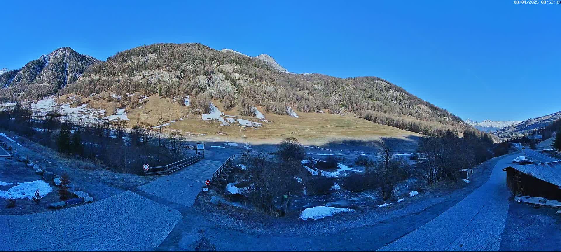 A mountain valley with a small wooden bridge, patches of snow, and a gravel road under a clear blue sky.