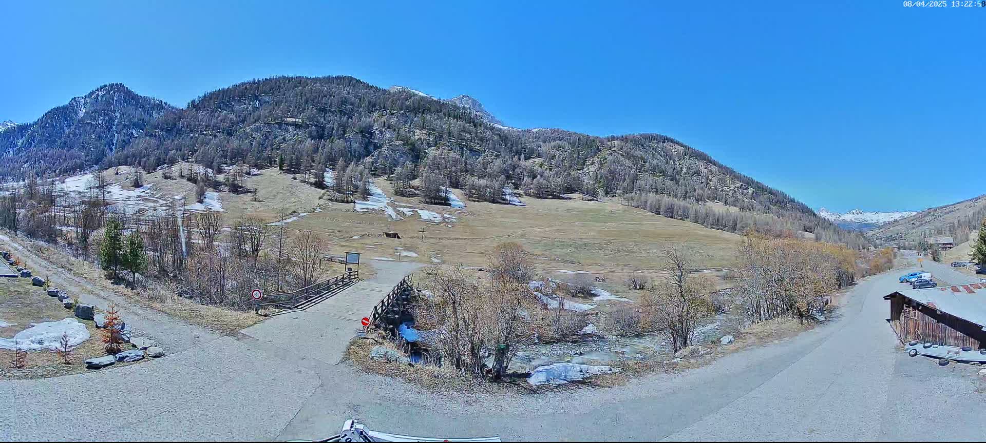A mountain valley scene on a sunny day shows a gravel road leading to a wooden bridge over a small stream, with patches of snow remaining on the hillside and budding trees lining the valley floor.