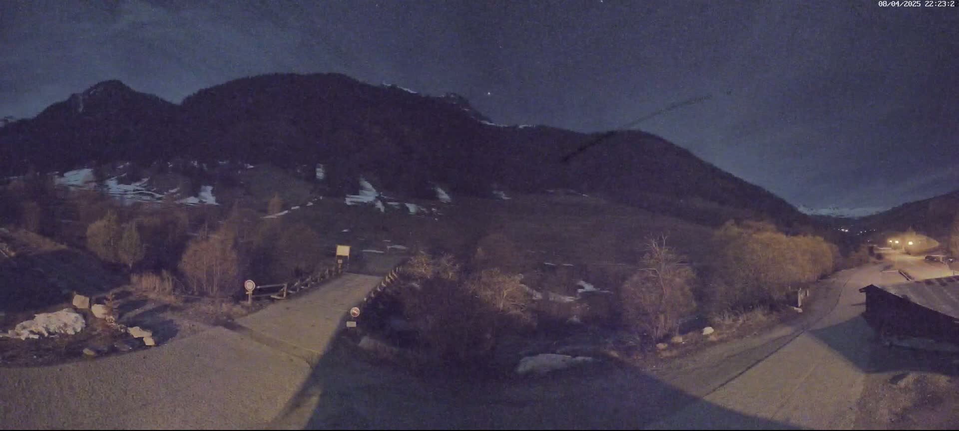 A nighttime view of a small bridge leading to a road that winds through a valley under a dark sky, with a mountain range in the background and some patches of snow visible.