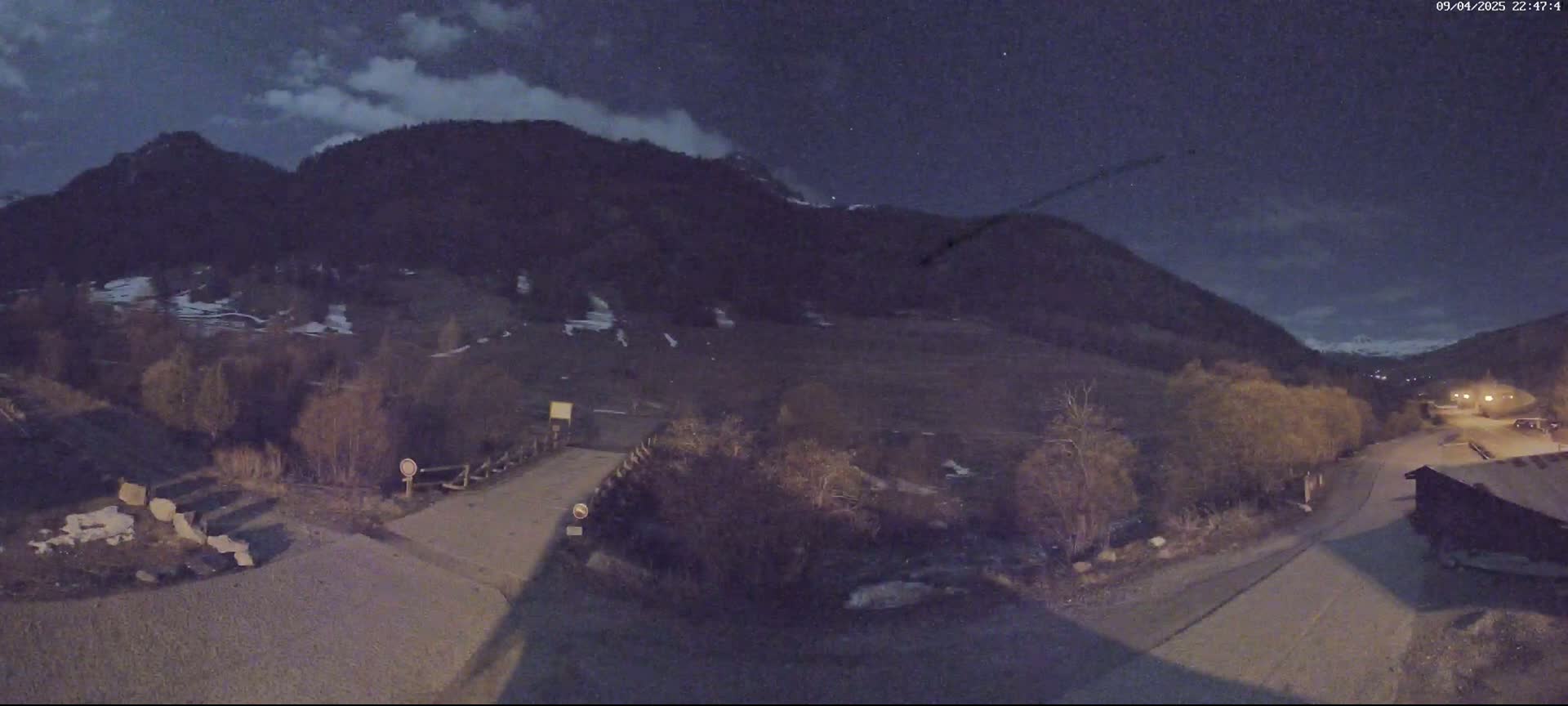 A nighttime view of a mountain valley under a partly cloudy sky, showing a road leading to a small building near a bridge.