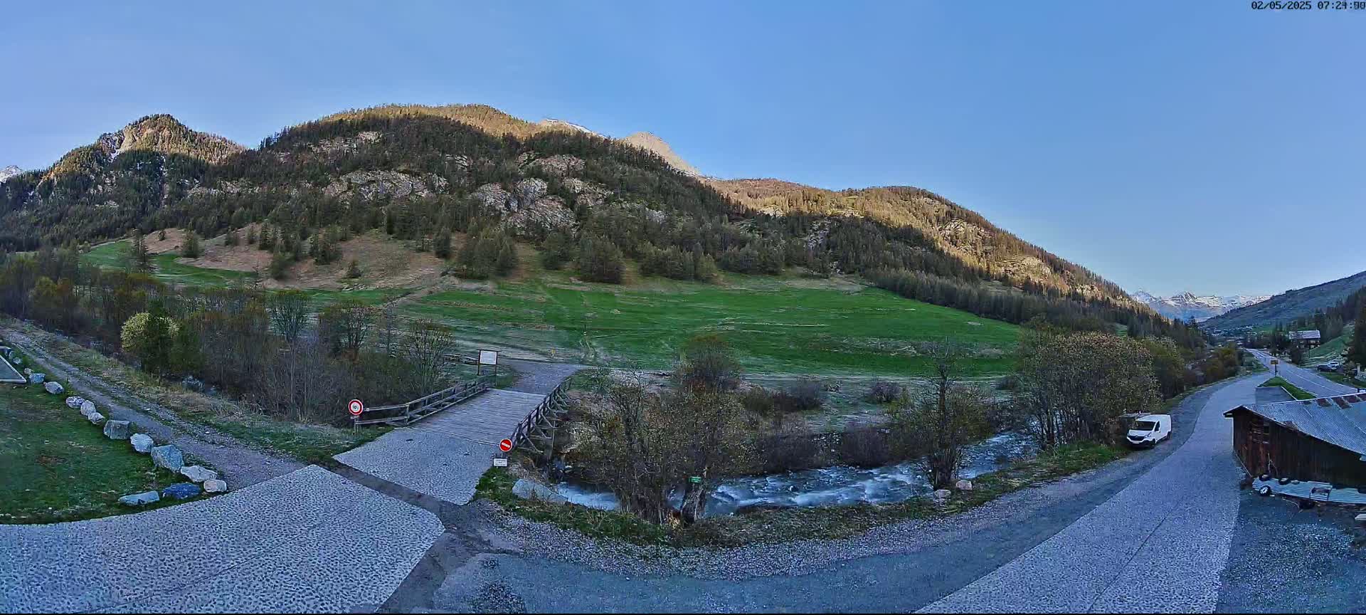A small wooden bridge crosses a stream in a valley nestled between mountains under a clear blue sky.