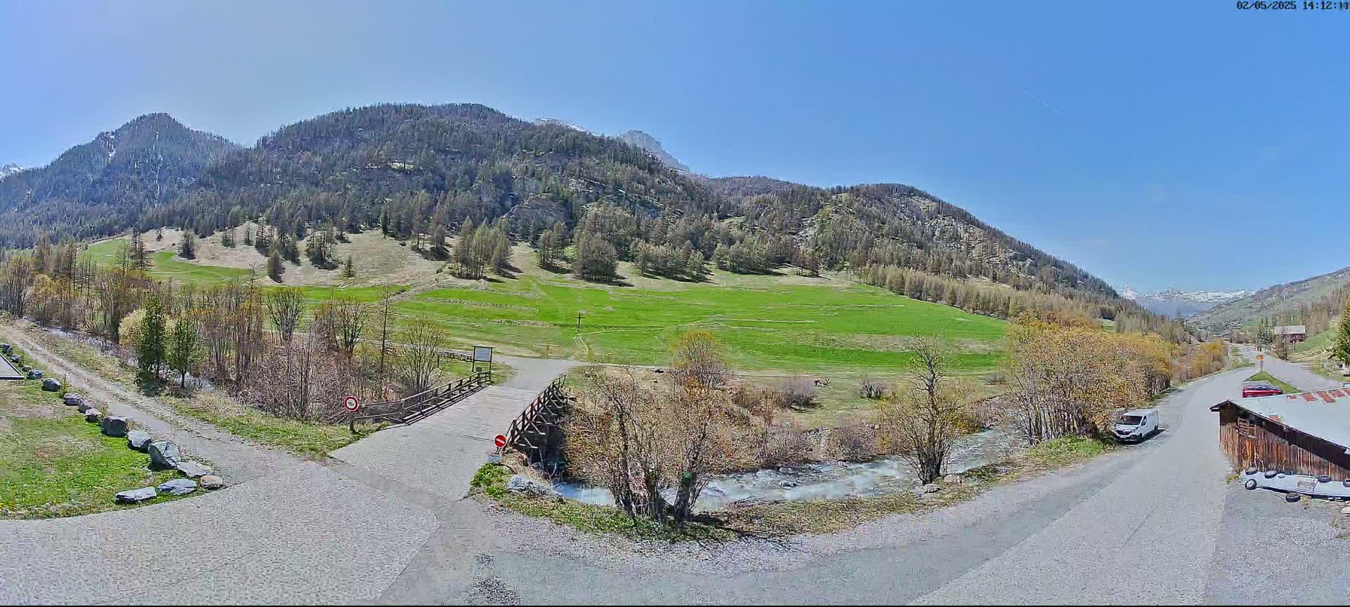A small wooden bridge crosses a stream in a valley surrounded by green fields and mountains under a clear blue sky.