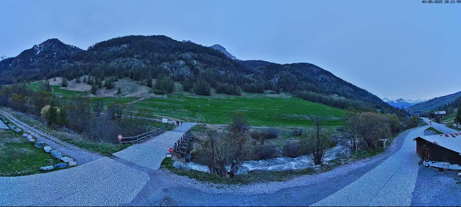 A wooden bridge crosses a small stream in a valley nestled between mountains under a twilight sky.