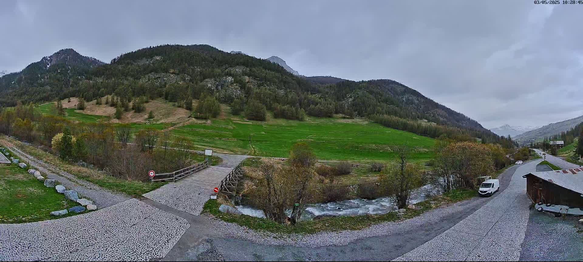 A wooden bridge crosses a small river in a valley surrounded by green hills and mountains under an overcast sky.