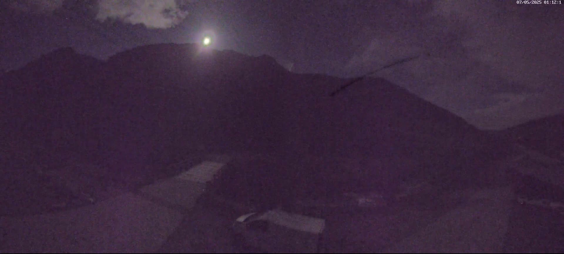 A nighttime, low-light view shows dark, silhouetted mountains under a partly cloudy sky with a bright light source near the peak of a mountain.