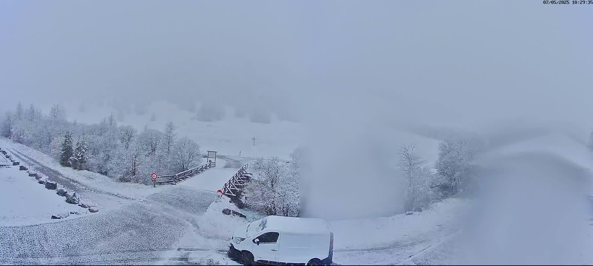 A snow-covered landscape with a white van parked near a snow-covered road and bridge, under heavy snowfall and foggy conditions.