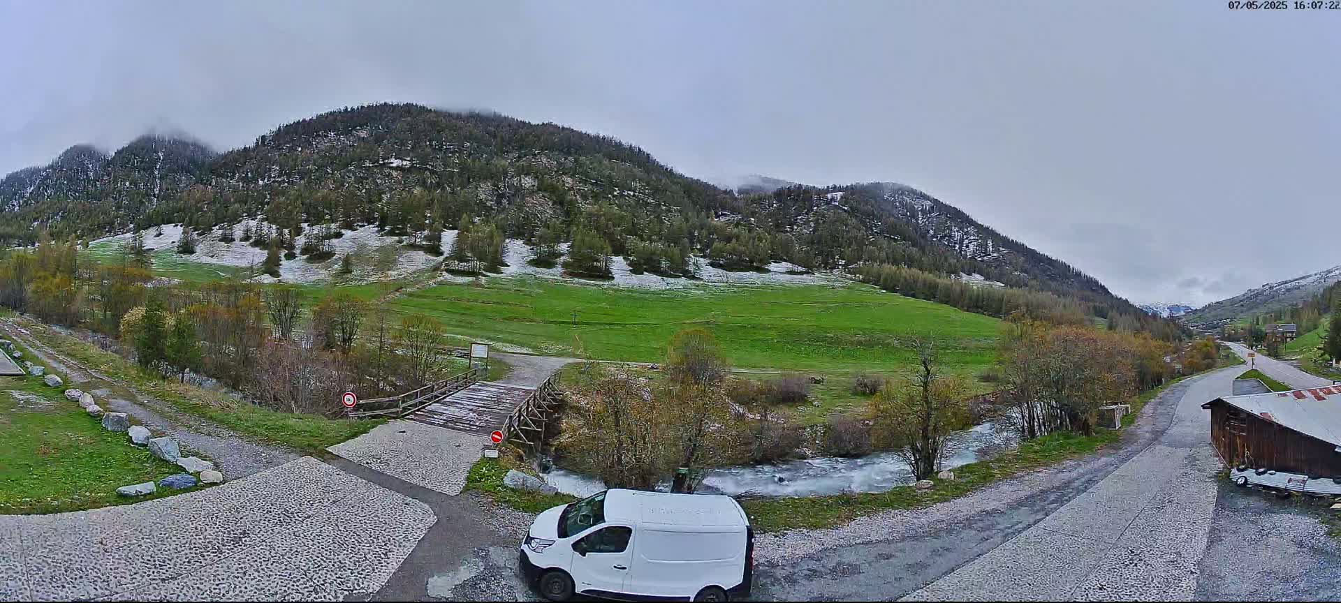 A white van is parked near a small wooden bridge over a stream, with a green valley and snow-dusted mountains in the background under an overcast sky.