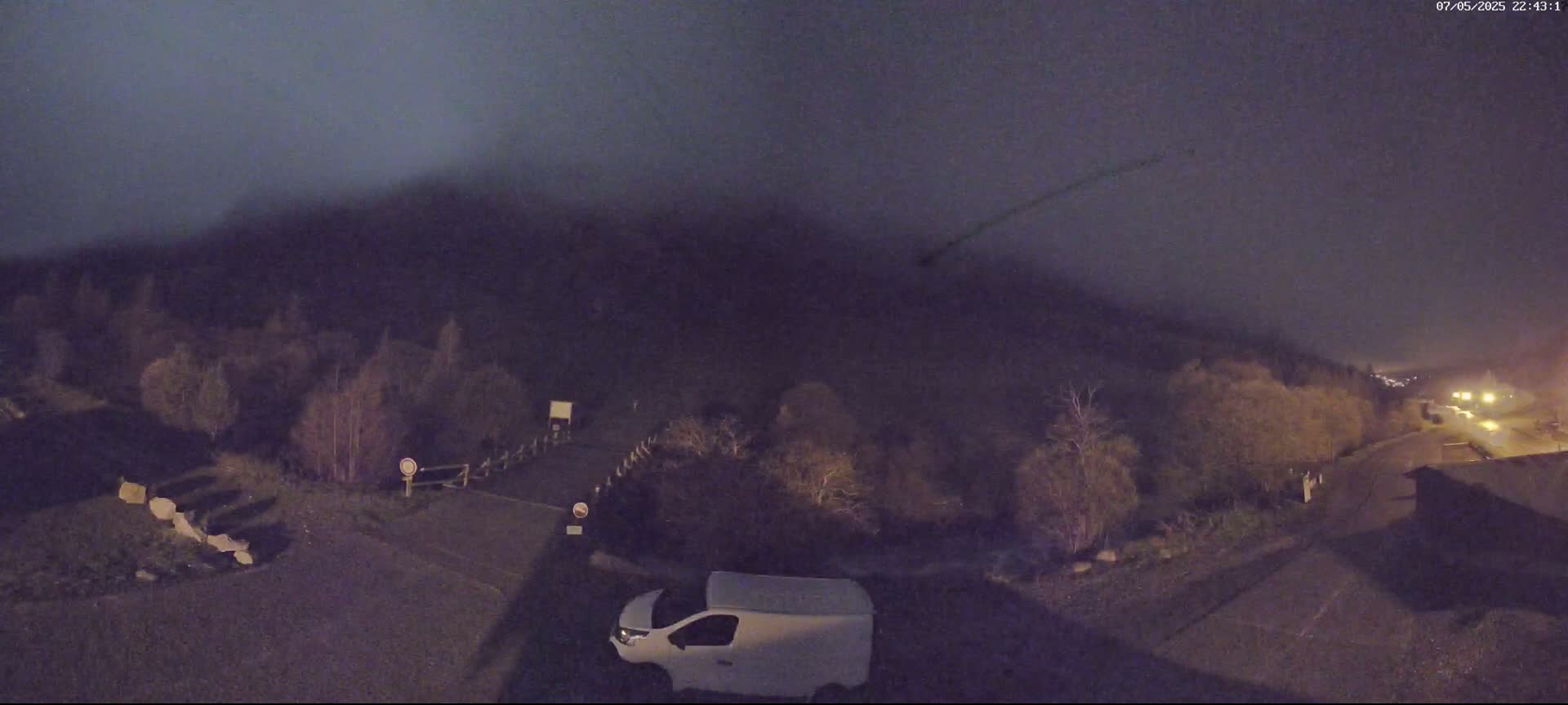 A nighttime, foggy scene shows a white van parked in a lot near a road, with trees and a hill in the background and a faint contrail in the sky.
