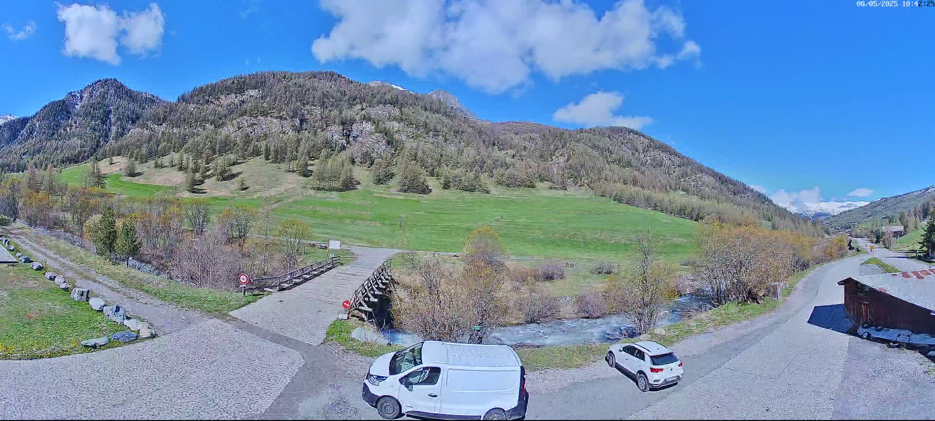 A sunny day in a mountainous region shows a valley with green fields, a small stream, a wooden bridge, two parked cars, and a van.