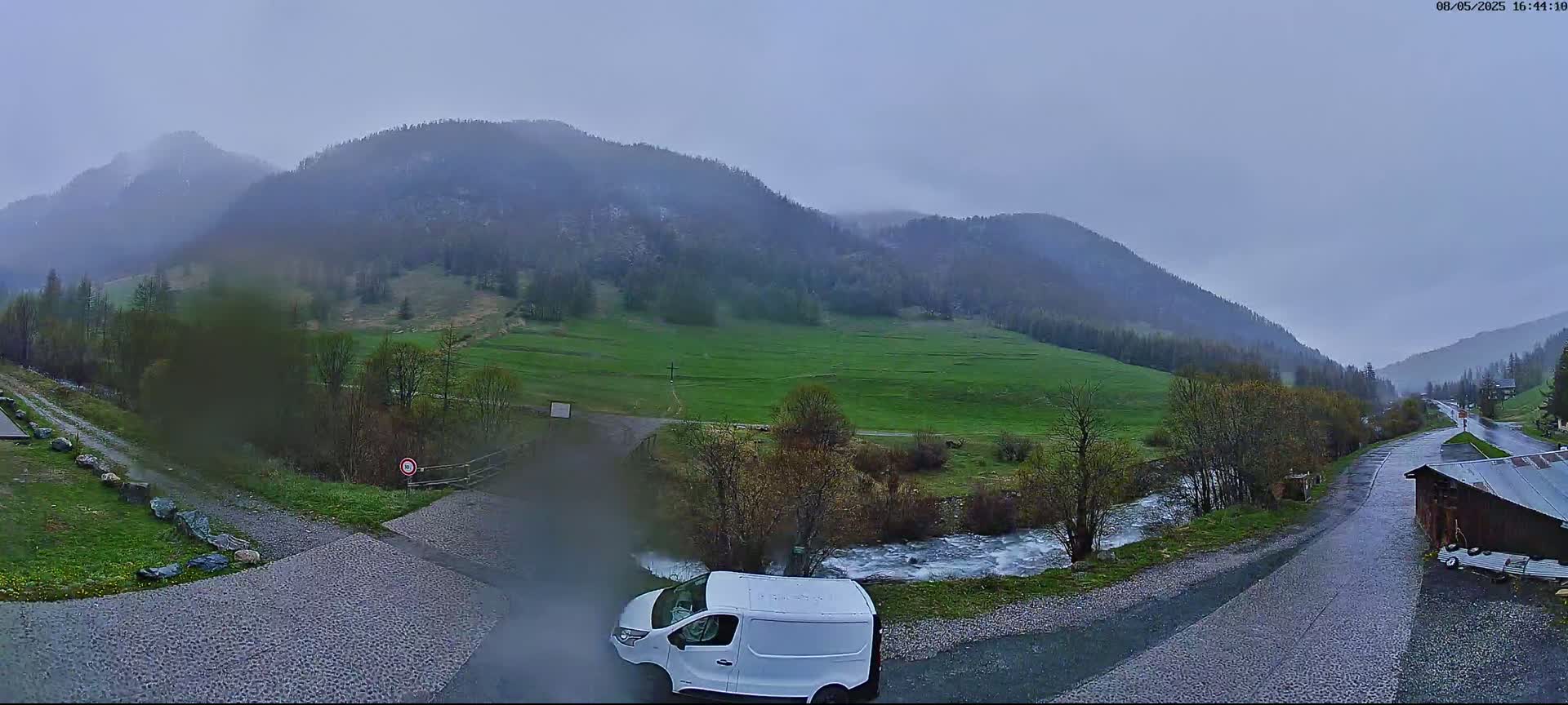A white van is parked near a fork in a wet road leading to a valley surrounded by misty, green mountains under an overcast sky.