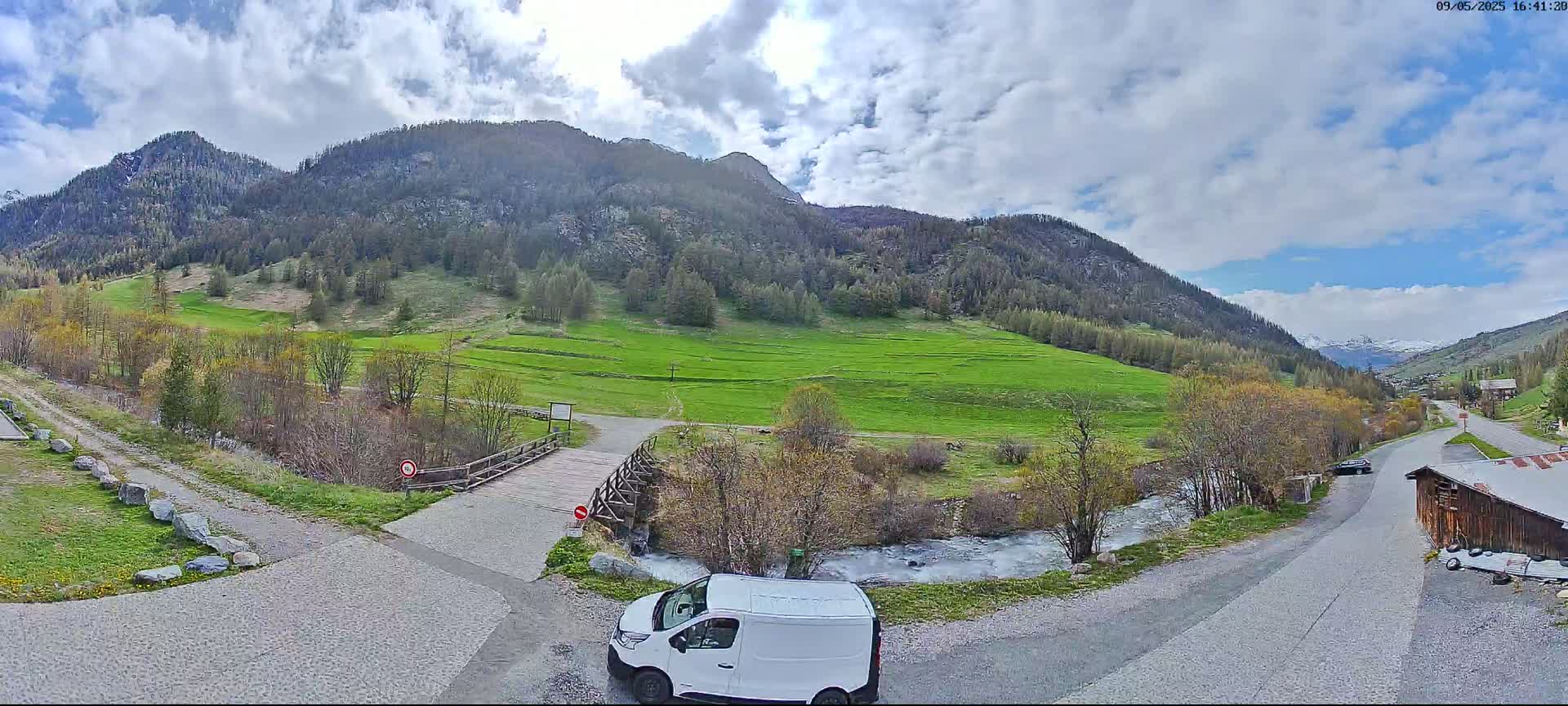 A partly cloudy day in a mountainous valley shows a white van parked near a small wooden bridge over a creek, with green fields and a road leading to a small village in the distance.
