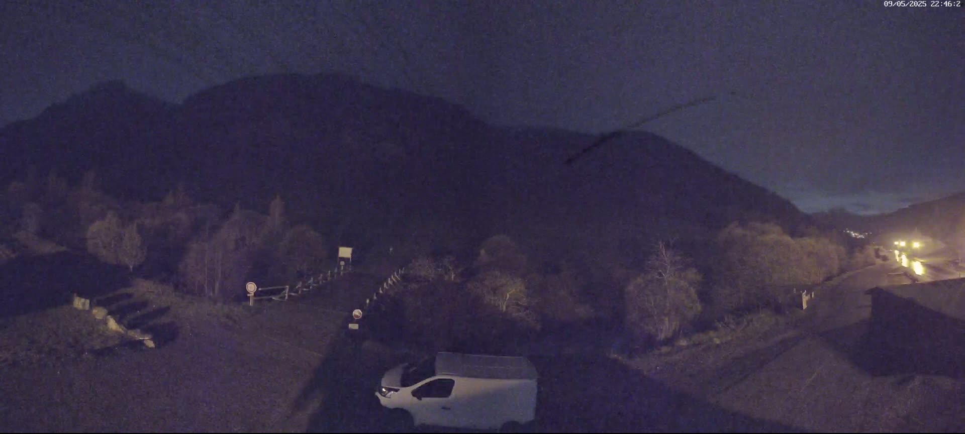 A white van is parked in a nighttime outdoor area near a road with a dark mountain in the background under a dark sky.