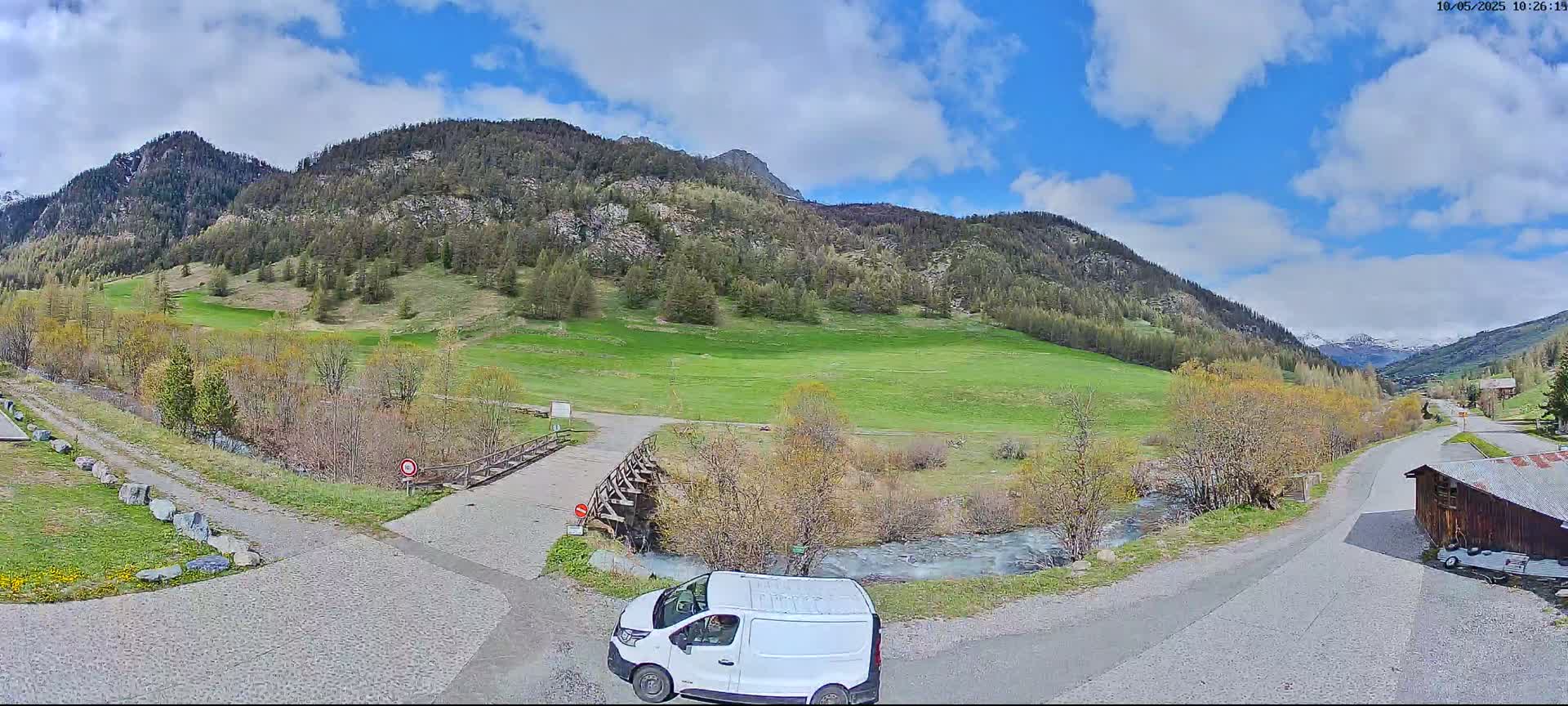 A white van is parked near a paved road that leads to a wooden bridge over a small stream, with a green field and mountains in the background under a partly cloudy sky.