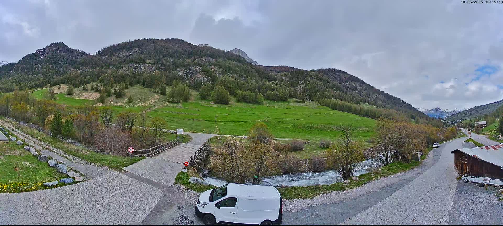 A white van is parked near a paved road that leads to a wooden bridge over a small stream, with green fields and mountains in the background under an overcast sky.
