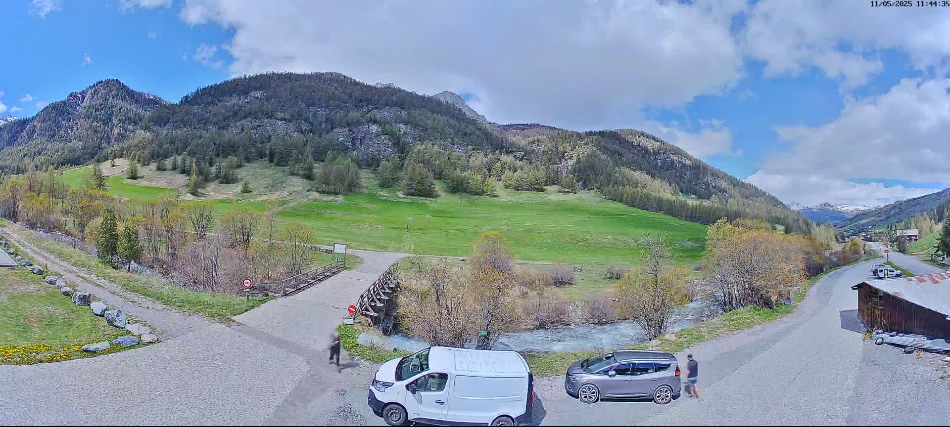 A partly cloudy day reveals a valley scene with green grass, trees, a small river, mountains in the background, and two parked vehicles near a person walking.