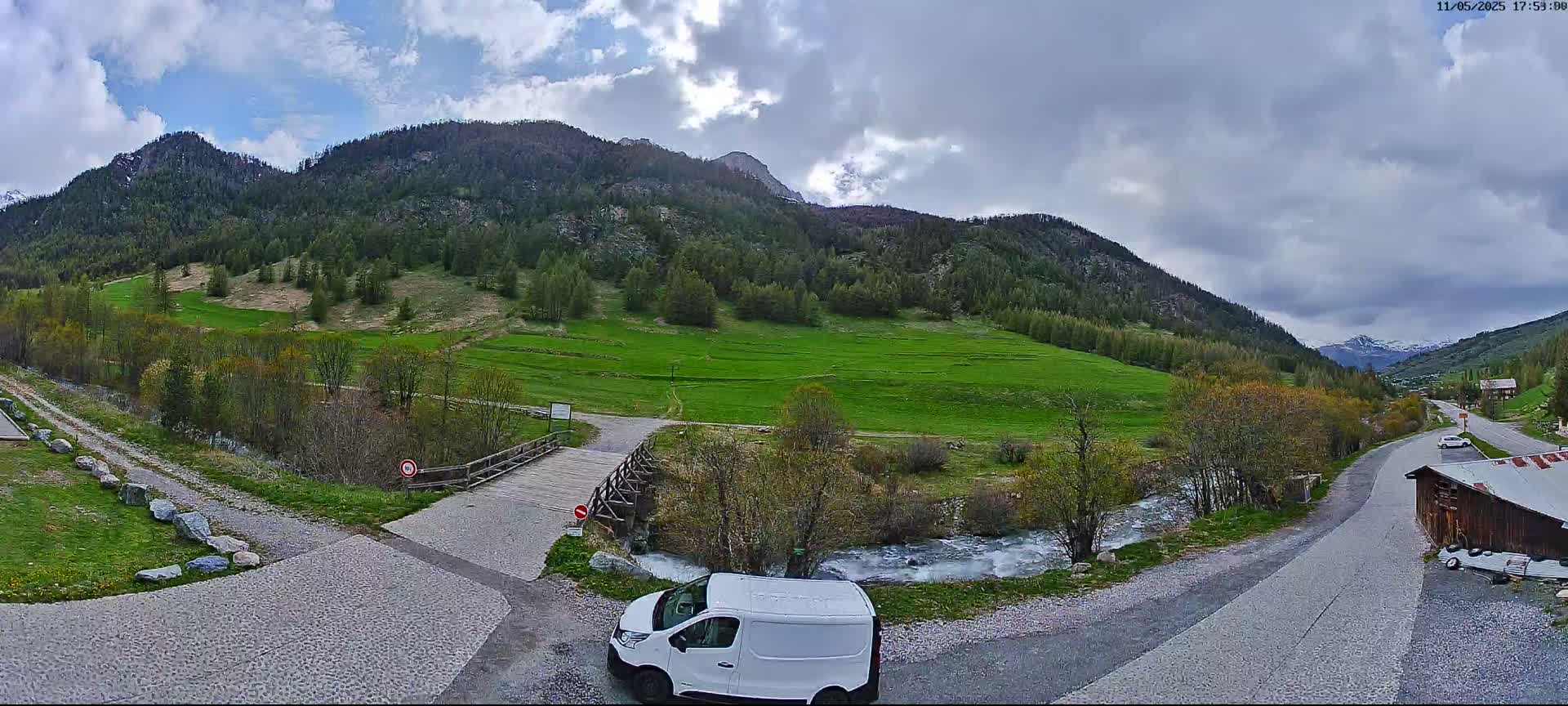 A white van is parked near a crossroads overlooking a valley with a small river, green fields, and mountains under a partly cloudy sky.
