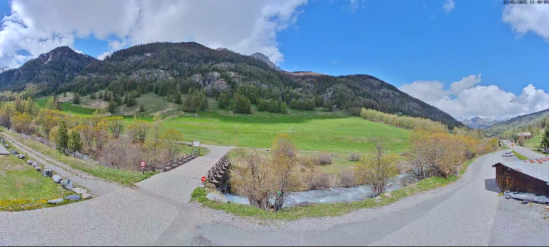 A partly cloudy sky overlooks a valley with green fields, a small stream, and a wooden bridge, with mountains in the background.