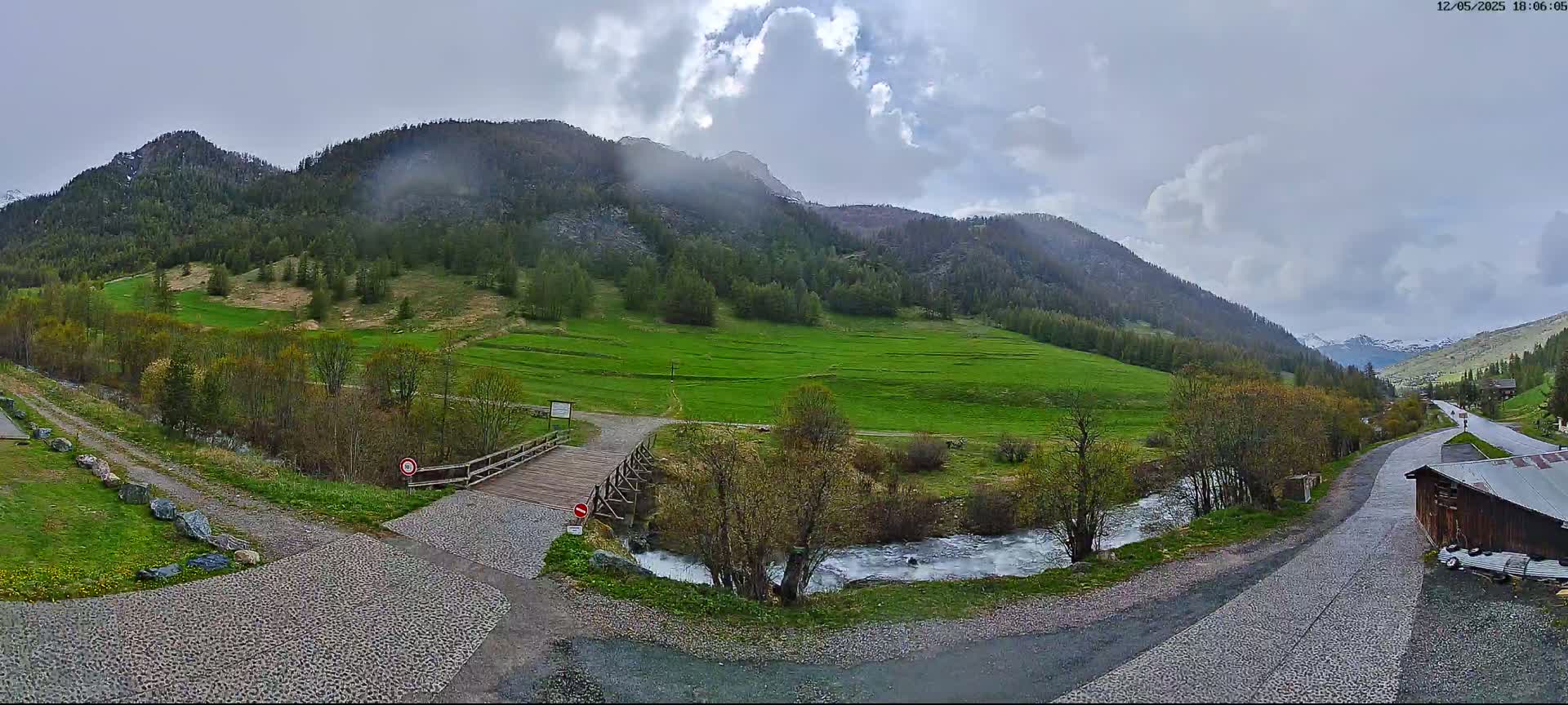A small wooden bridge crosses a stream in a green valley nestled between cloudy mountains.