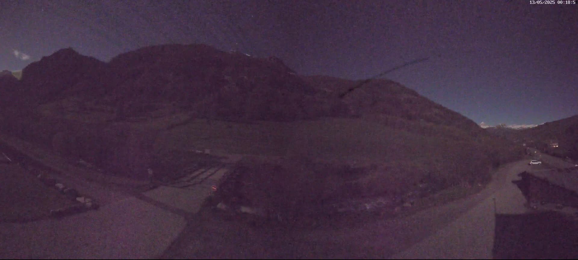 A nighttime view shows dark mountains under a mostly clear, dark sky with a few stars visible, a road, and some parked vehicles.