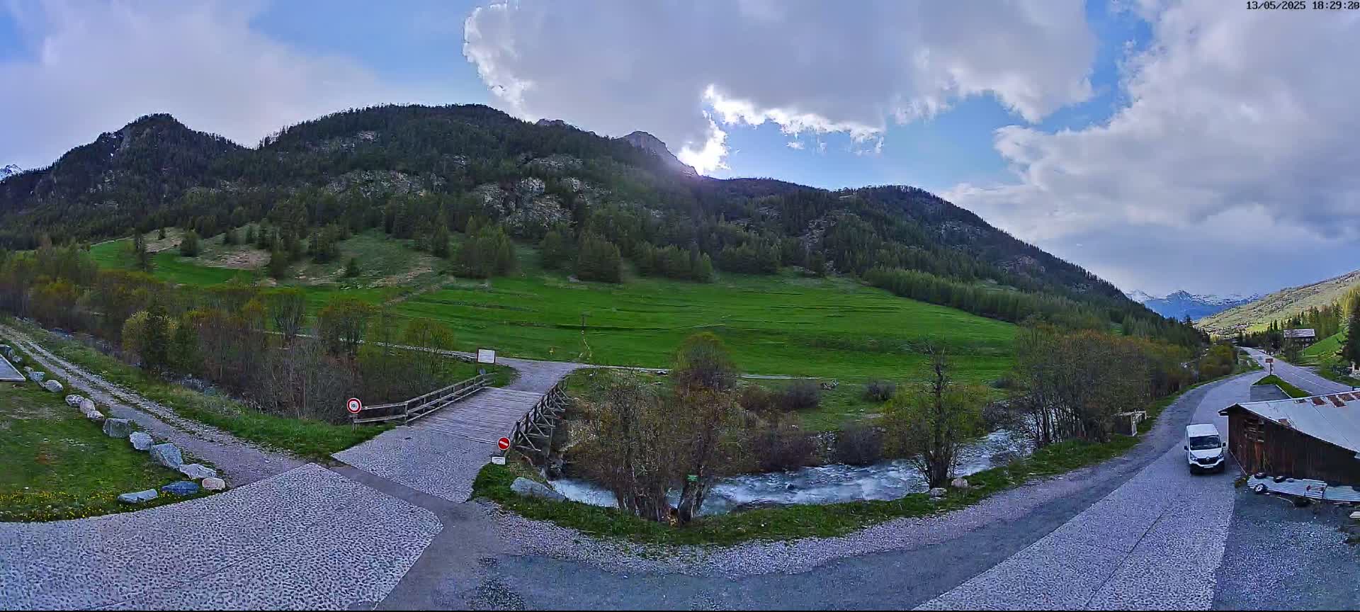 A partly cloudy sky overlooks a valley with a small wooden bridge over a stream, green fields, and mountains in the background.