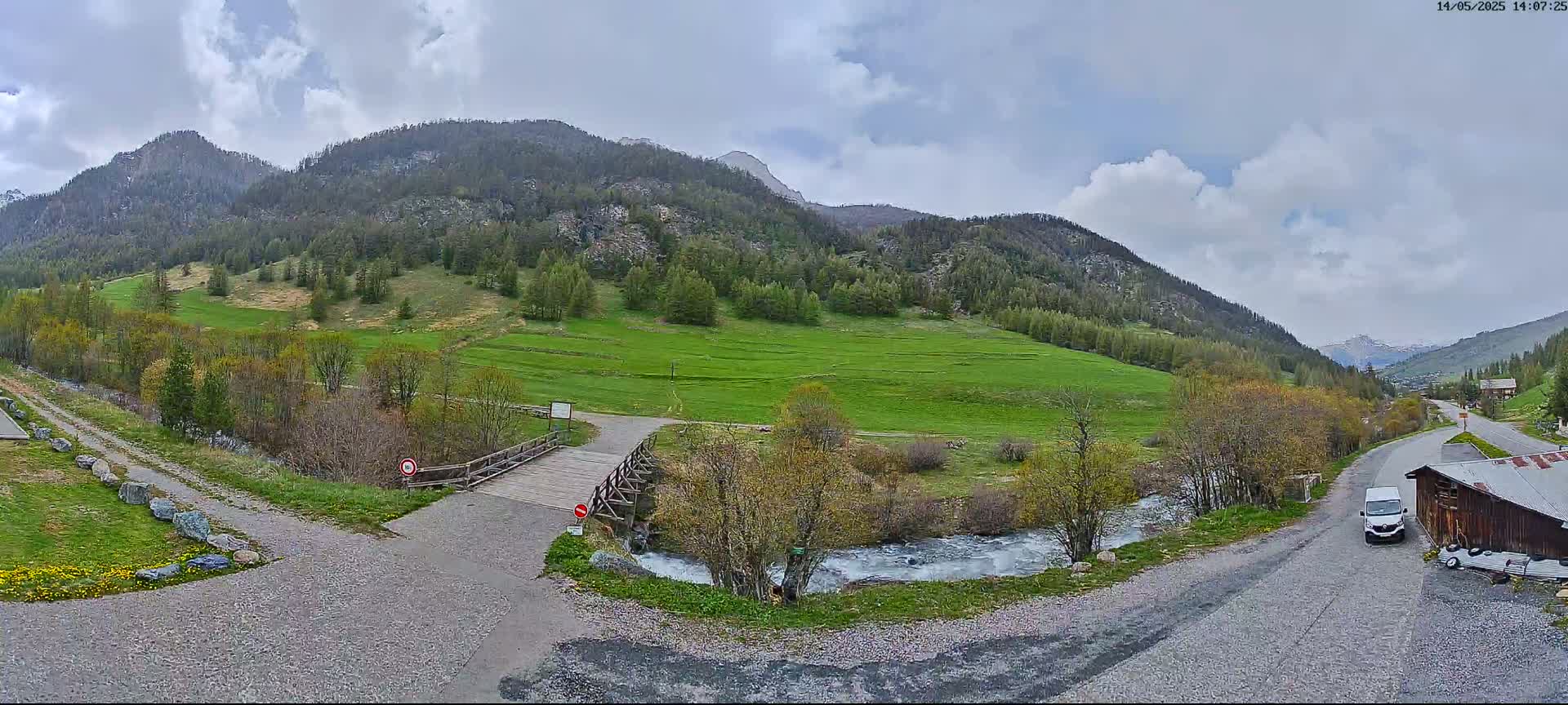 A mountain valley scene under a partly cloudy sky shows a small wooden bridge over a stream, roads leading to a building, and a van parked nearby.