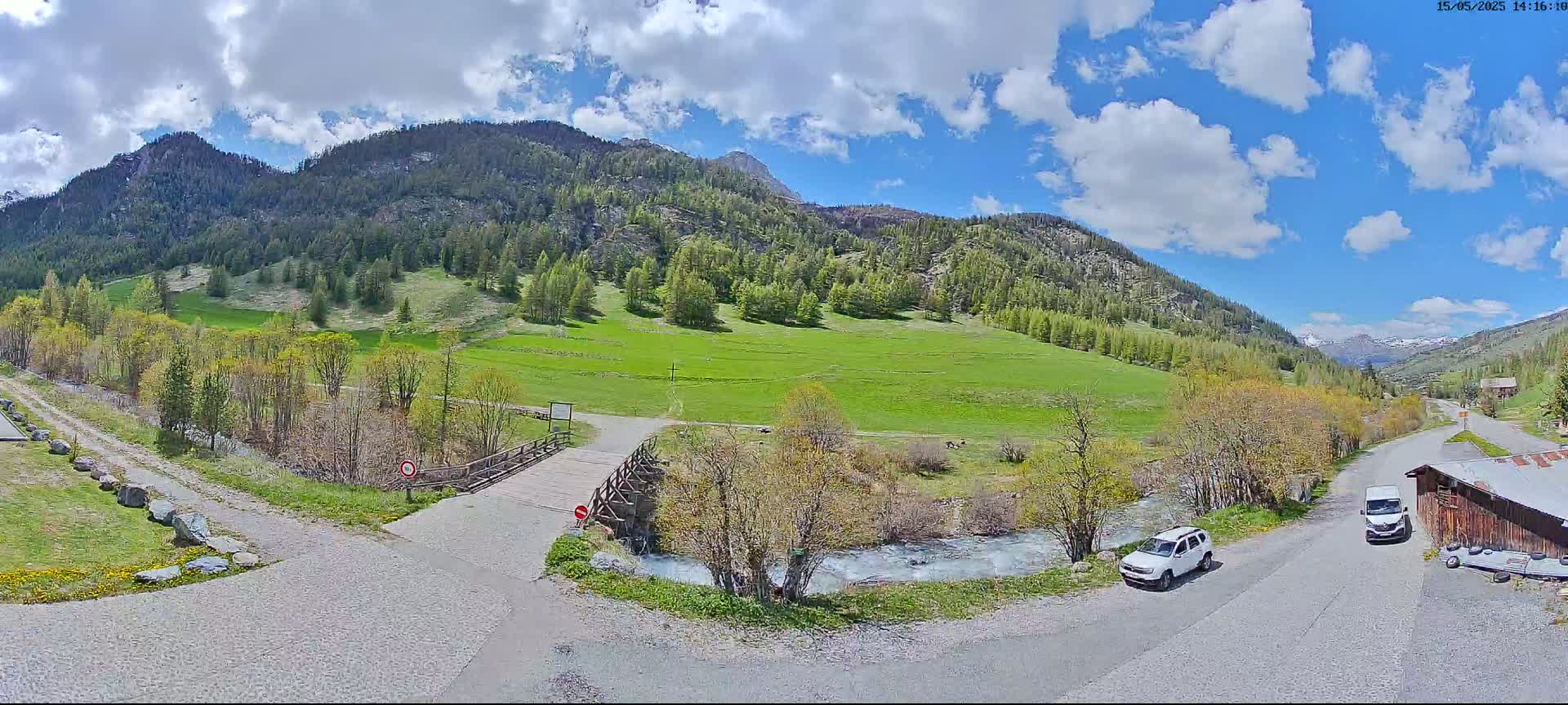 A partly cloudy day reveals a mountainous valley with a grassy field, a small river, a wooden bridge, and two parked vehicles near a building.