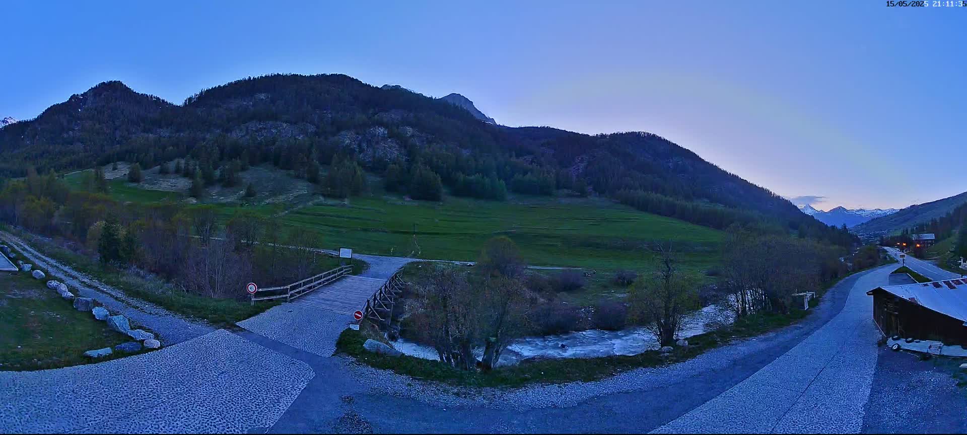 A wooden bridge crosses a small stream in a valley nestled between mountains under a twilight sky.