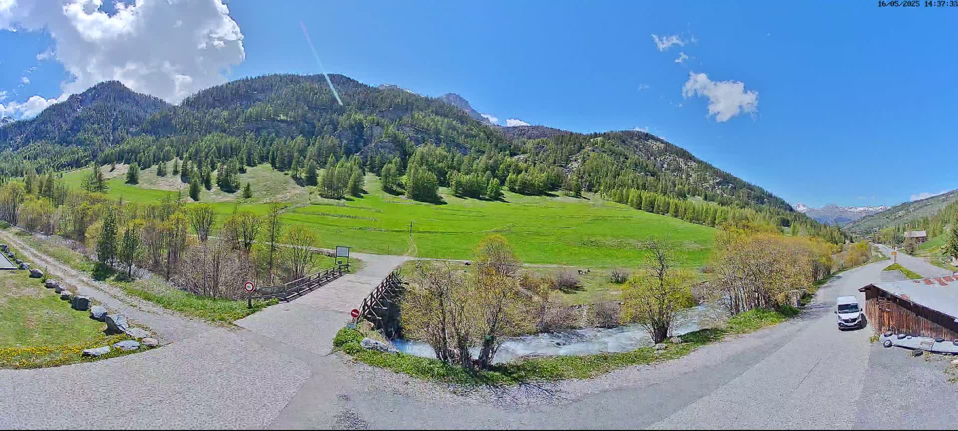 A sunny day in a mountainous valley shows a small bridge over a stream, leading to a grassy field surrounded by green trees and mountains, with a paved road and a van parked nearby.