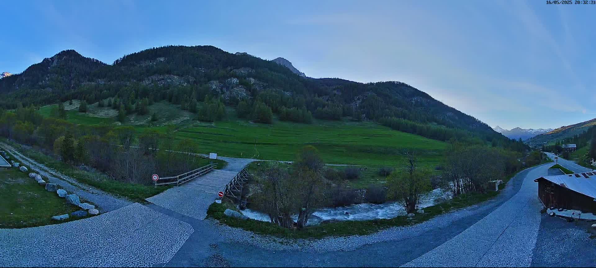 A wooden bridge crosses a small stream in a valley surrounded by green hills and mountains under a clear blue sky.