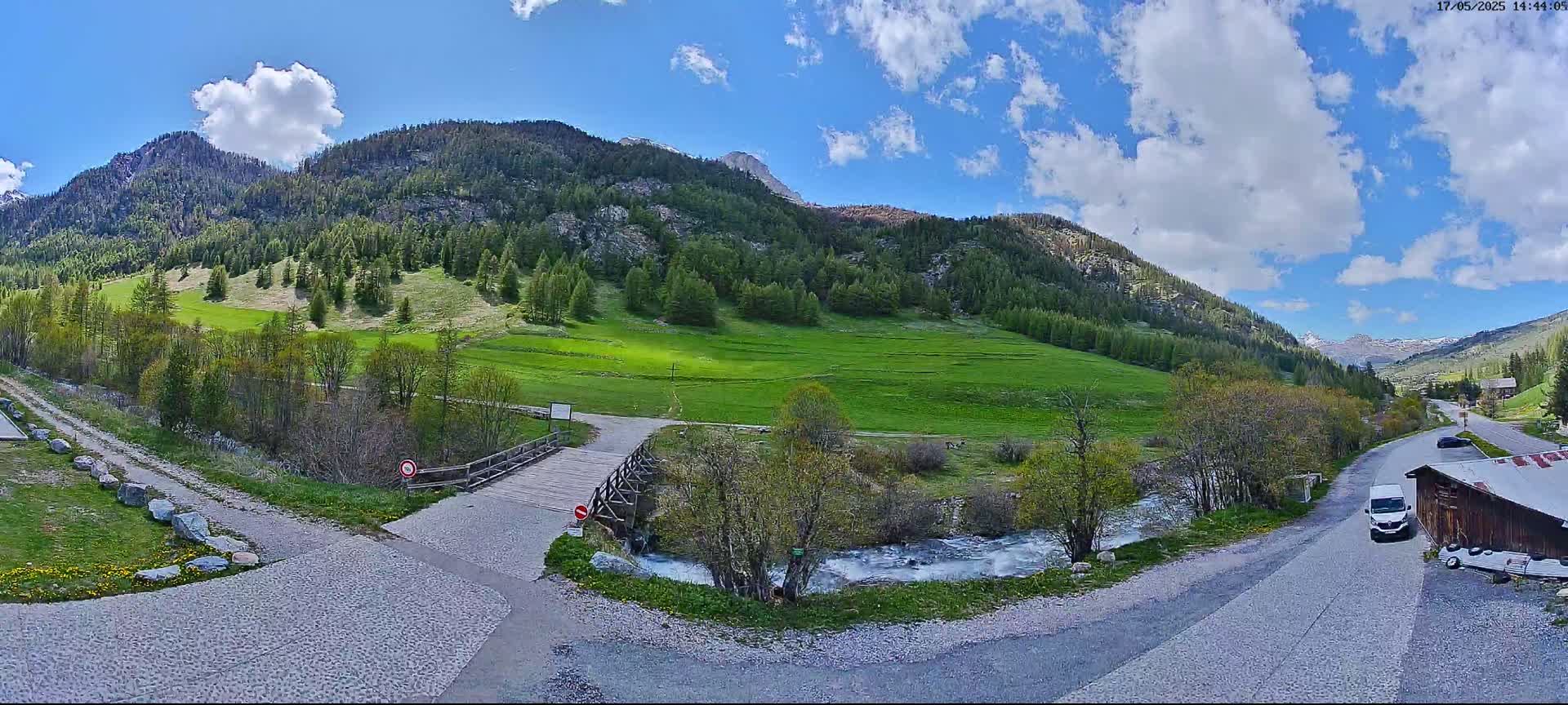 A wooden bridge crosses a small stream in a mountainous valley under a partly cloudy blue sky.