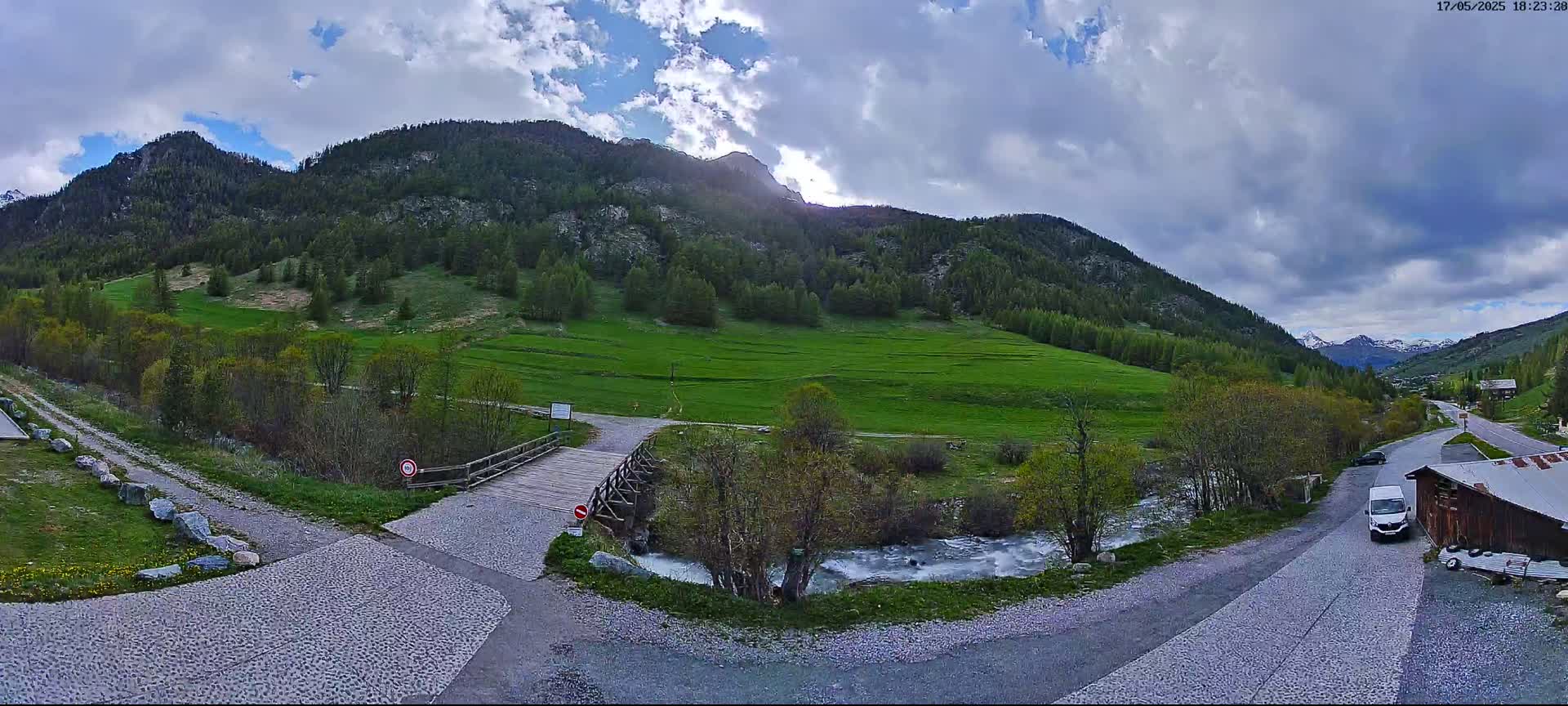 A wooden bridge crosses a small river in a valley surrounded by green hills and mountains under a partly cloudy sky.