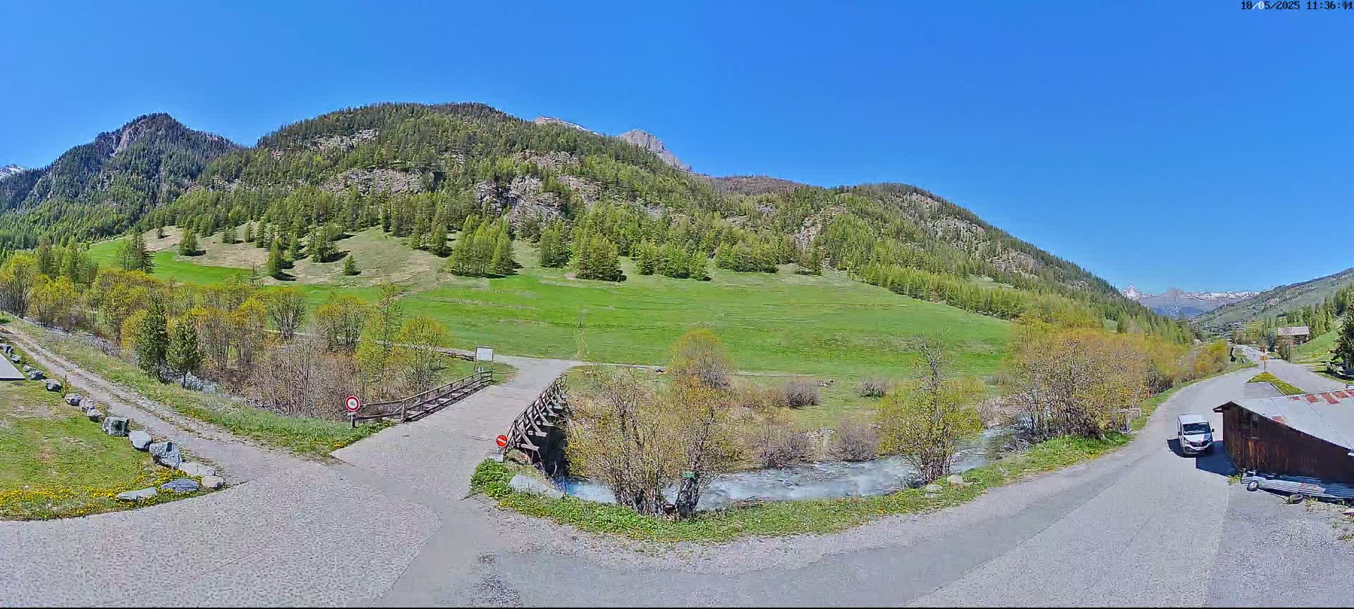 A paved road forks near a wooden bridge over a small stream, with a grassy hillside and coniferous-covered mountains in the background under a clear blue sky.