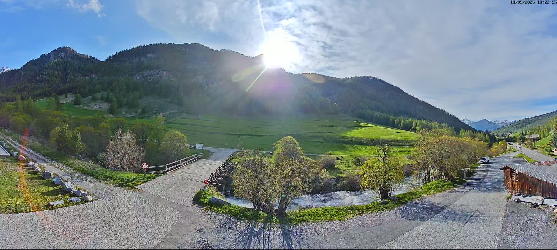 A sunny day in a mountainous valley shows a wooden bridge over a river, with green fields and trees, and a roadway leading to a building.