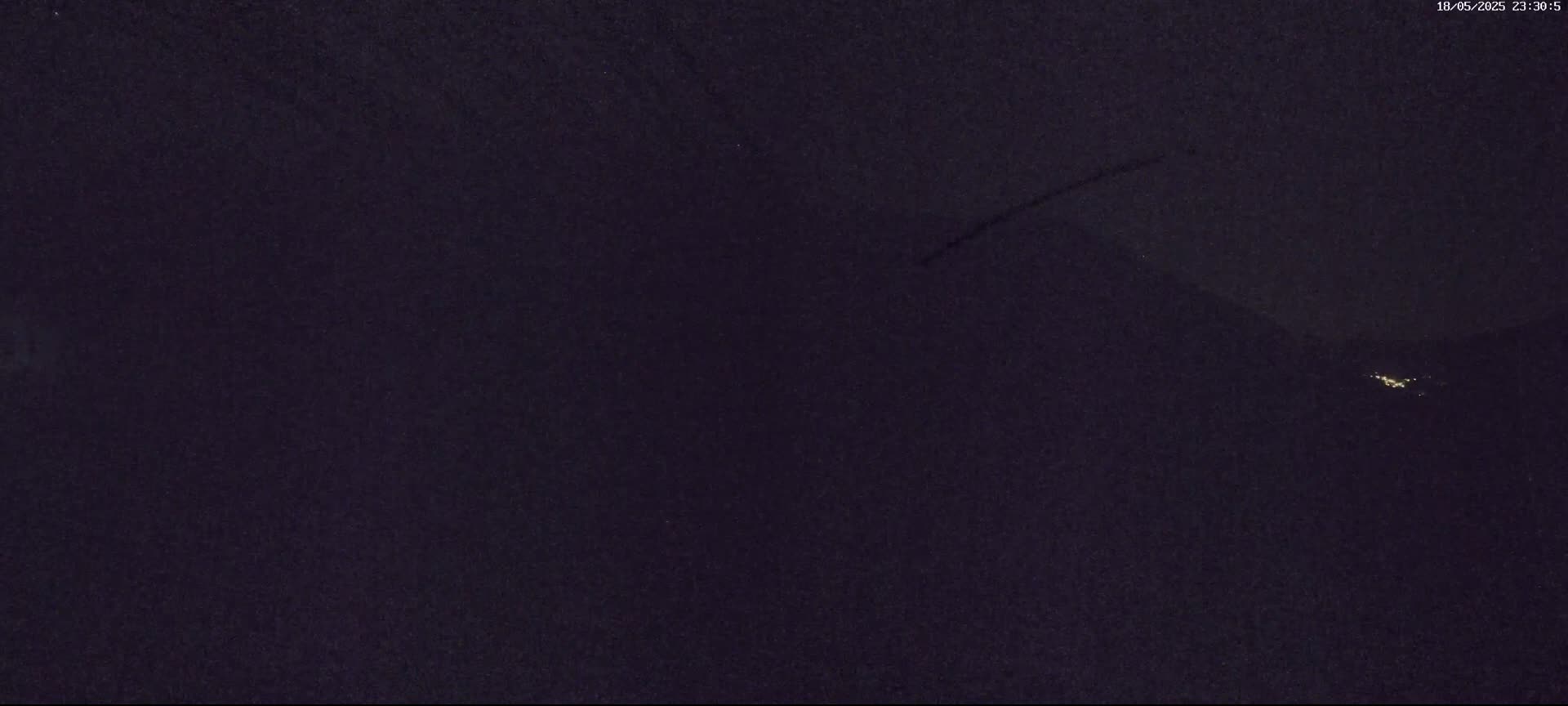 A nighttime, dark image shows a faint contrail in the sky above a distant cluster of lights on a dark landscape.